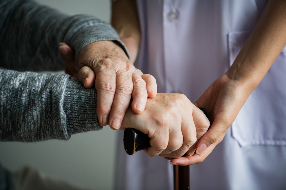 Nahaufnahme von Händen, die einen Gehstock halten, eine jüngere Person hält unterstützend die Hand eines älteren Menschen